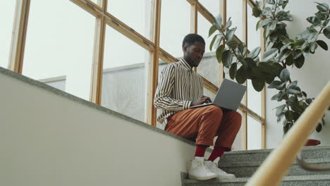 Afro-Mann-Sitzt-In-Einem-Modernen-Gebäude-Und-Arbeitet-Am-Laptop